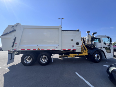 Mack LR64 refuse truck in side view showing rear compactor, hydraulic arm, and white cab with chrome wheels.