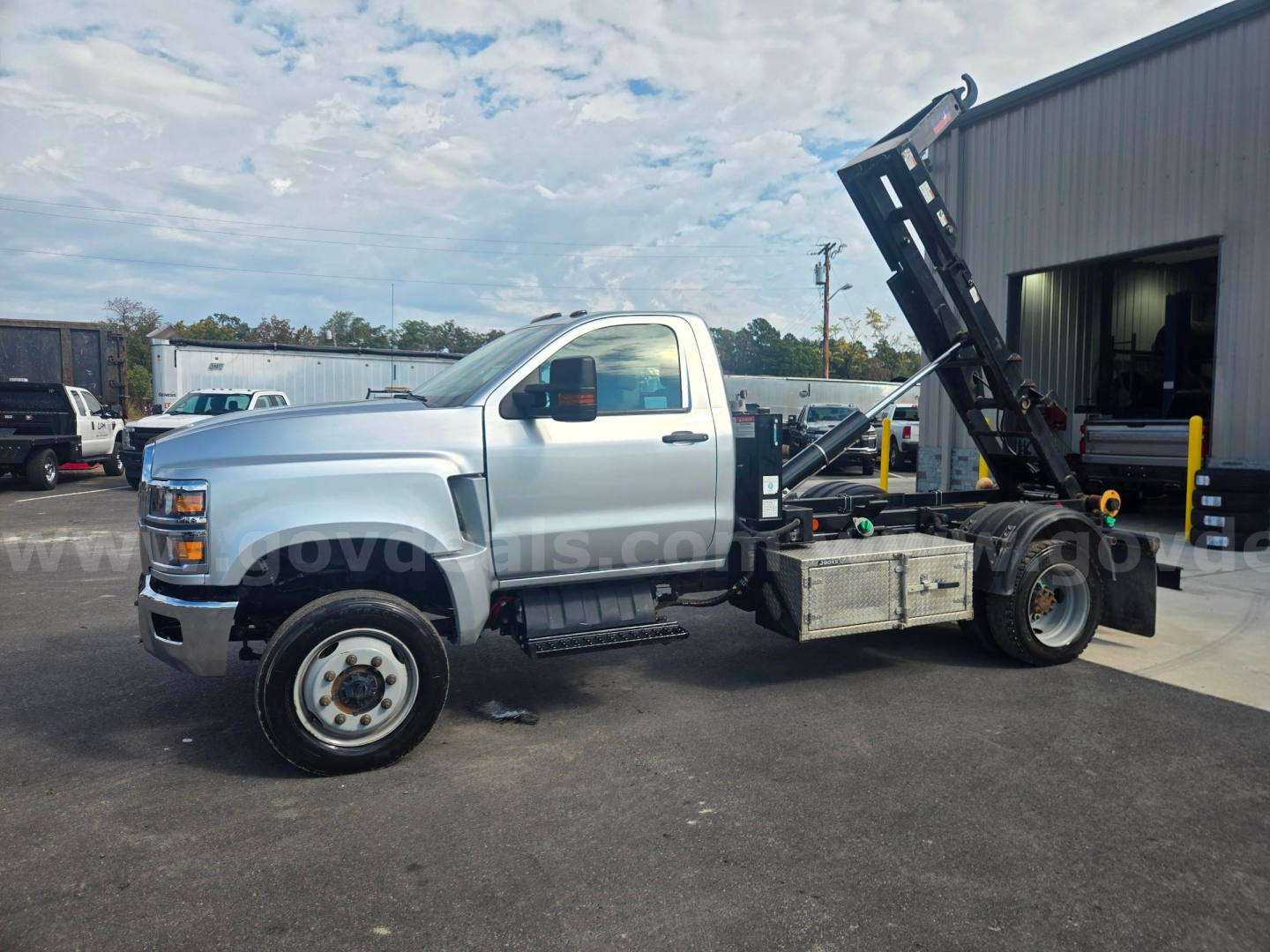 Truck bed with hydraulic hooklift on a Chevrolet 6500 platform showing utility toolbox, diesel engine cab, and wheel. Visible text on truck not legible.