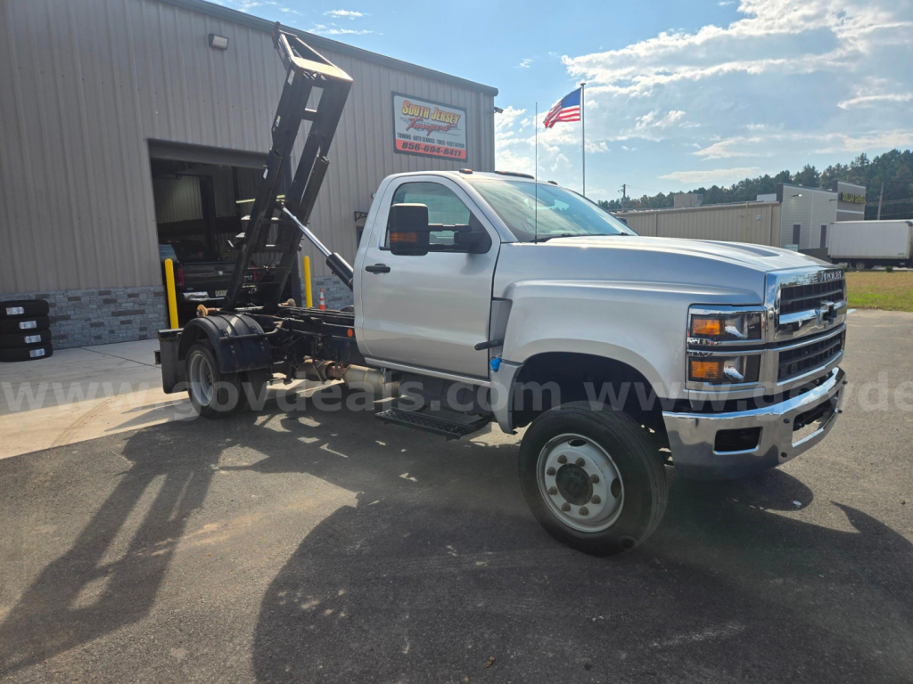 Chevrolet 6500 dump truck chassis in side profile with tipping hoist and front chrome bumper, white cab, DOT tires, dealership lot in background.