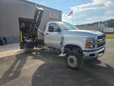 Chevrolet 6500 dump truck chassis in side profile with tipping hoist and front chrome bumper, white cab, DOT tires, dealership lot in background.