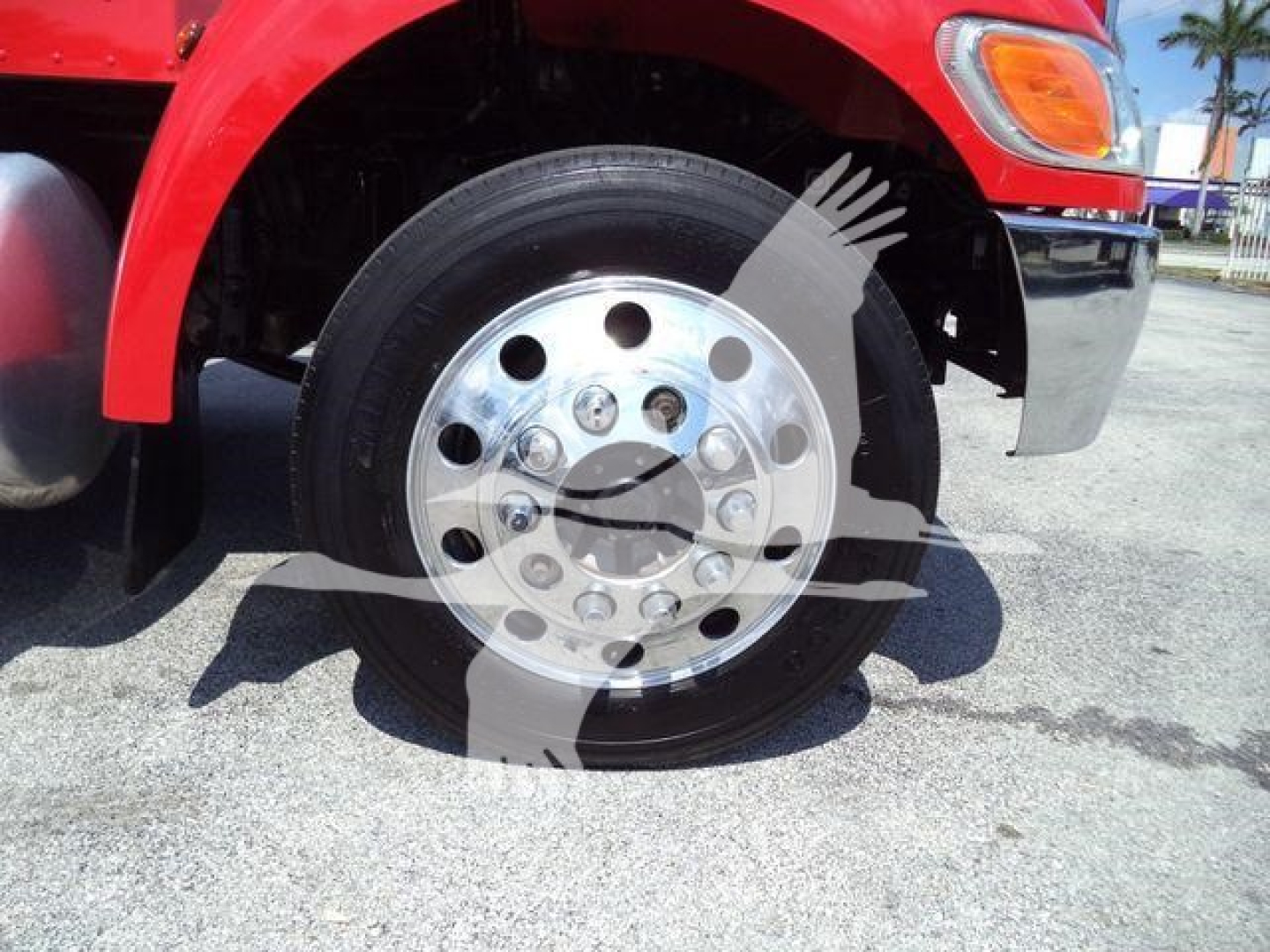 Front wheel of red Peterbilt truck with chrome hubcap and multiple lug nuts, partial fender and headlight visible.