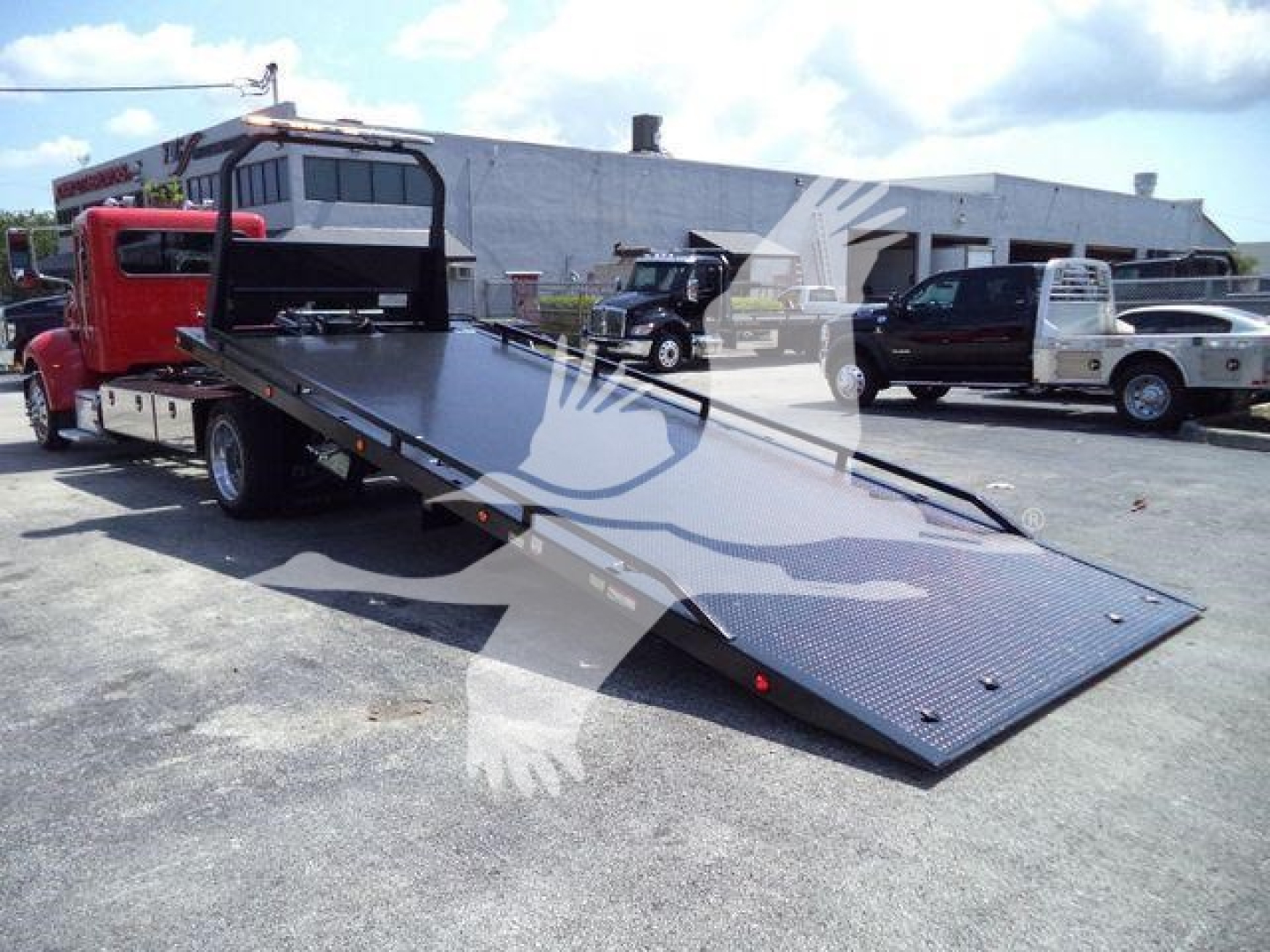 Peterbilt 337 rollback truck with a lowered flatbed ramp extended, red cab, other trucks in background.