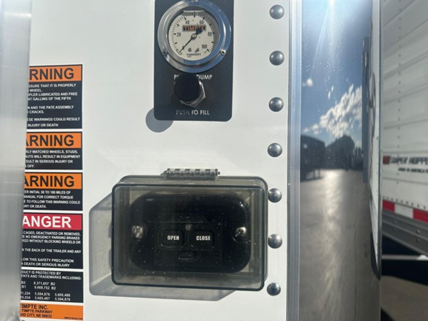 Timpte hopper trailer control panel and warning labels on white exterior with gauge and toggle switch visible.