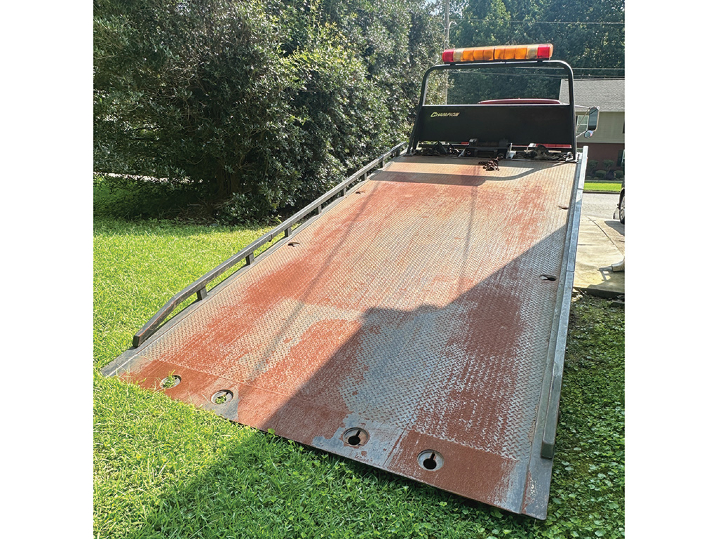 Truck bed of a 1999 Chevrolet 6500 rollback with rusted steel platform, rear ramps, and emergency light bar on cab; visible sunlit lawn and trees in background.