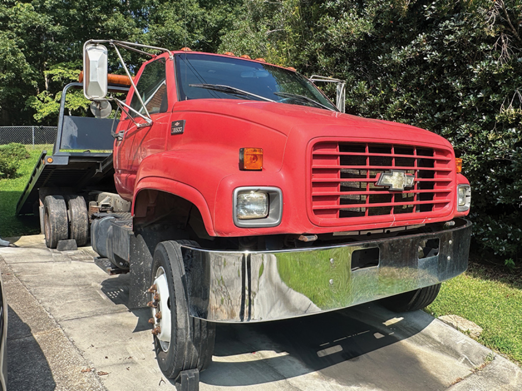 Truck front view of a red Chevrolet 6500 rollback wrecker with chrome front bumper and tilted cab, large side mirrors, and a tow bed in background.