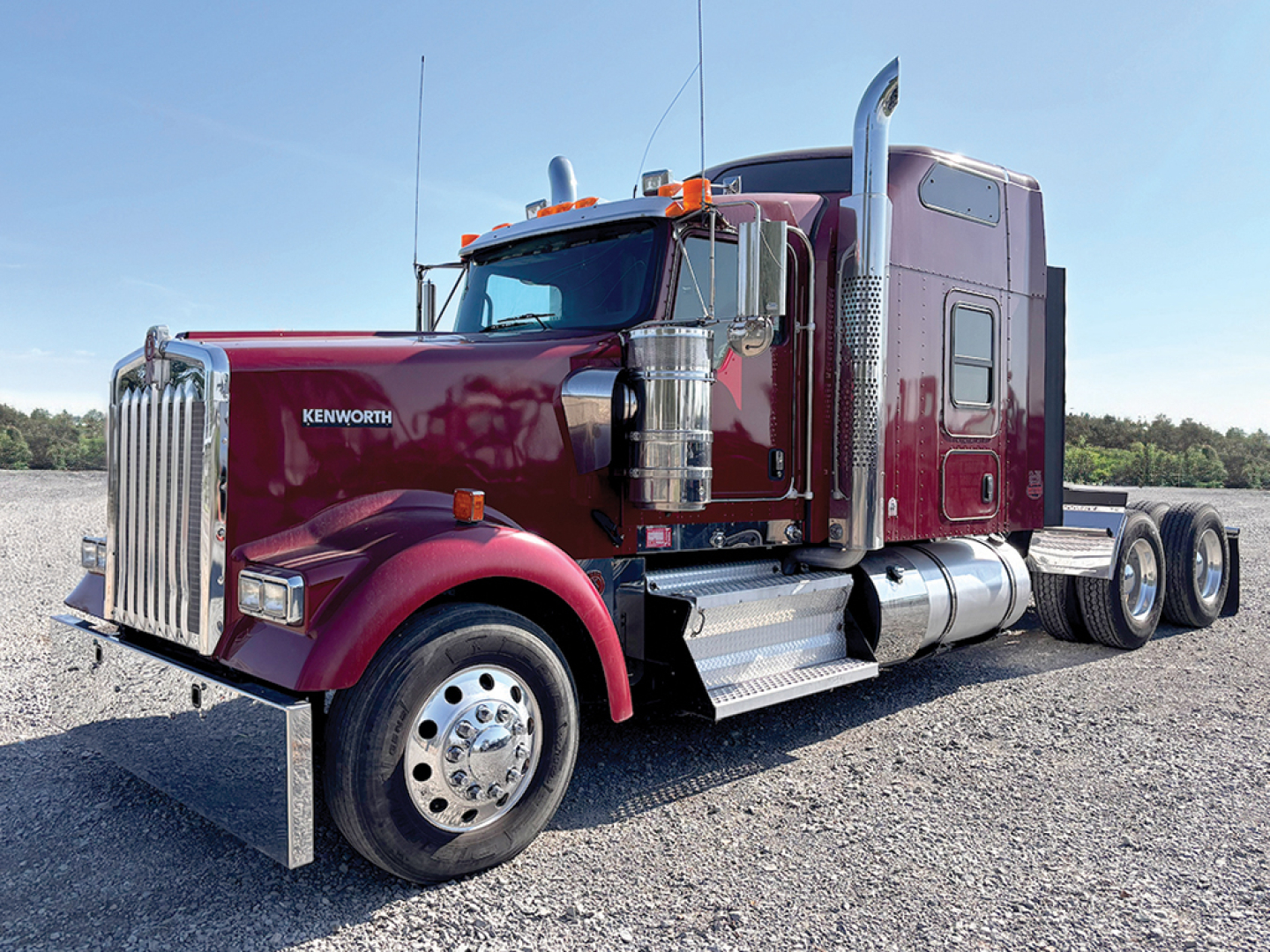Kenworth W-900L conventional sleeper truck in burgundy, front-left view, chrome exhaust stacks, dual rear axles, daytime sun glare.