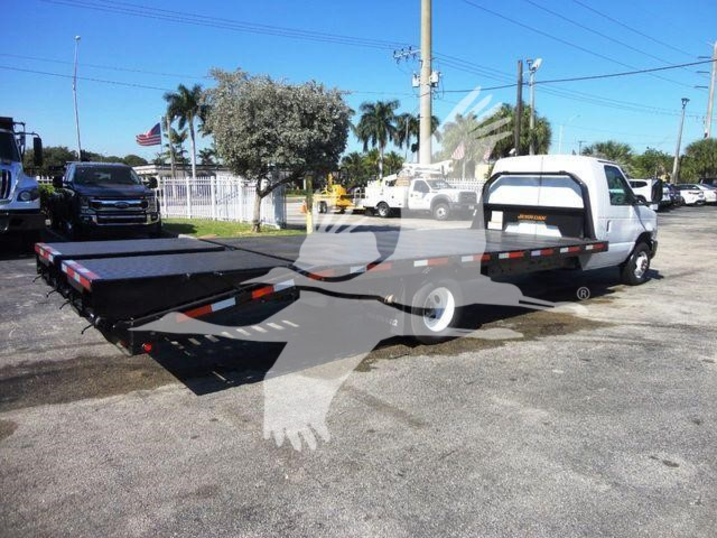 Flatbed tow truck with extended flatbed in yard. White Ford E450 cab; black flatbed with red/white reflective stripes; various vehicles in background; stock number visible on listing but not on image.