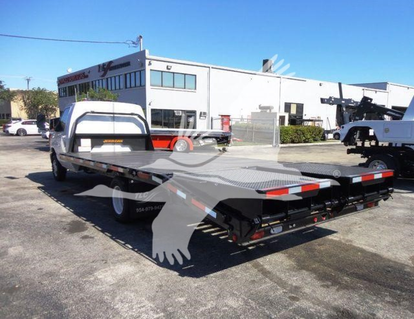 Flatbed tow truck chassis view showing a long black flatbed with a white cab in background, no legible text visible on side panels.