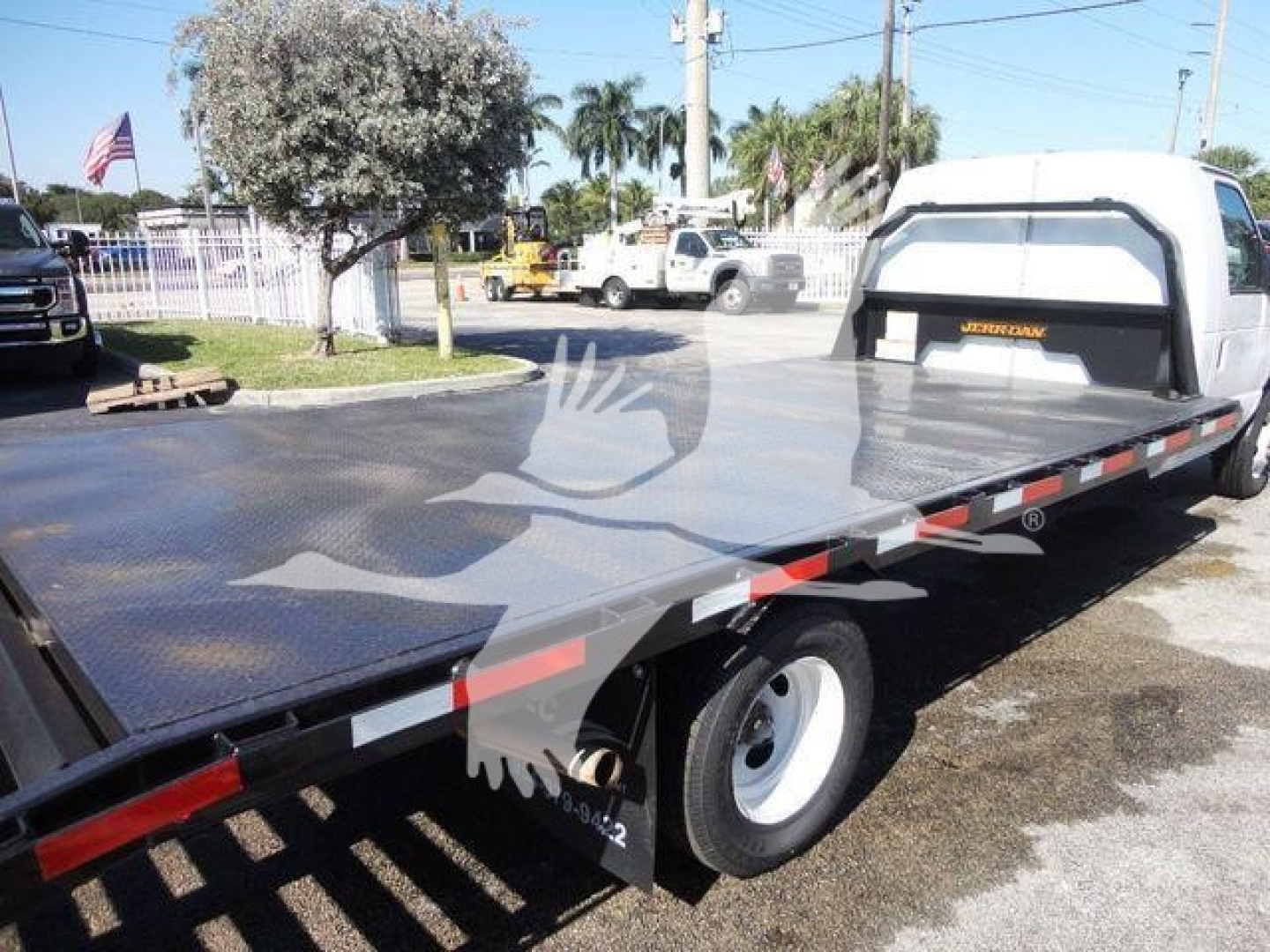 Ford E450 flatbed tow truck with long steel flatbed and white cab, rear wheels, and safety reflectors visible.