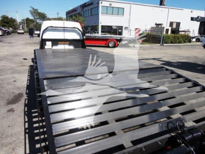 Flatbed truck bed attachment showing metal slats and wheel chocks with a Ford E-450 utility pickup in background.