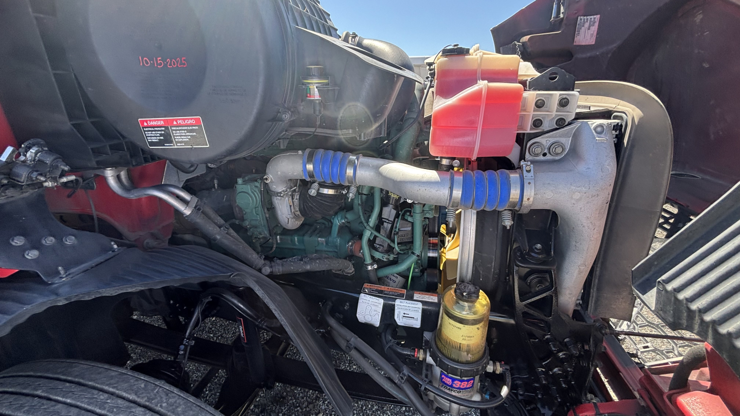 Volvo VNR engine bay showing label stickers, hoses, and fluid reservoirs; visible components include a black air reservoir, yellow oil filter, blue connected hoses, and red coolant reservoir cap.