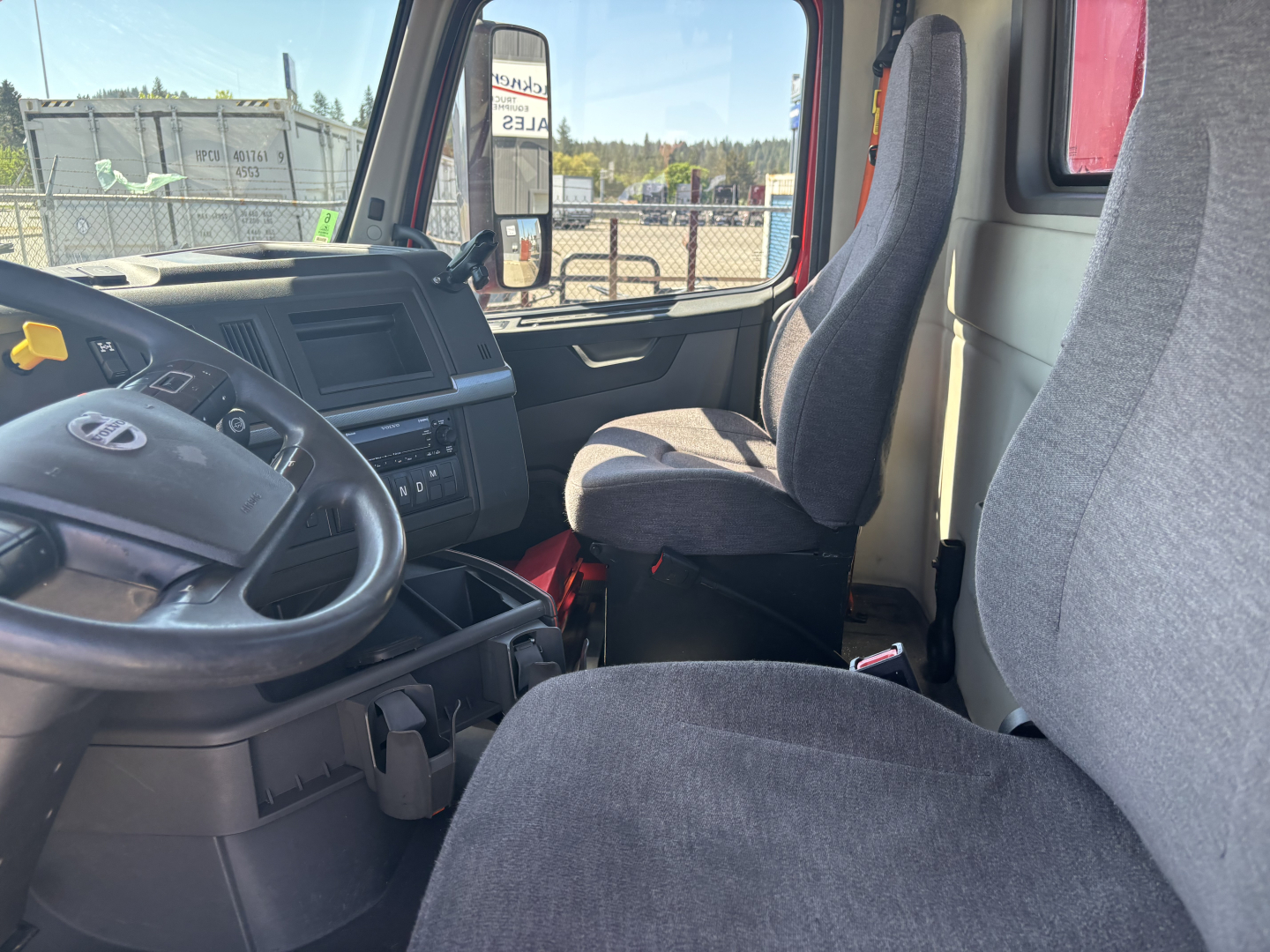Volvo VNR64T300 cab interior showing steering wheel dash and two front seats; gray fabric seats and black dashboard in daytime; side window view.