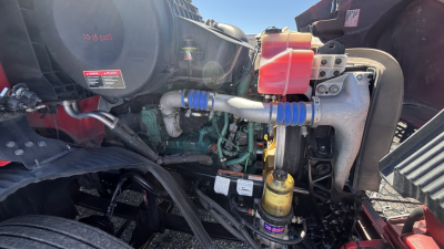 Volvo VNR engine bay showing label stickers, hoses, and fluid reservoirs; visible components include a black air reservoir, yellow oil filter, blue connected hoses, and red coolant reservoir cap.