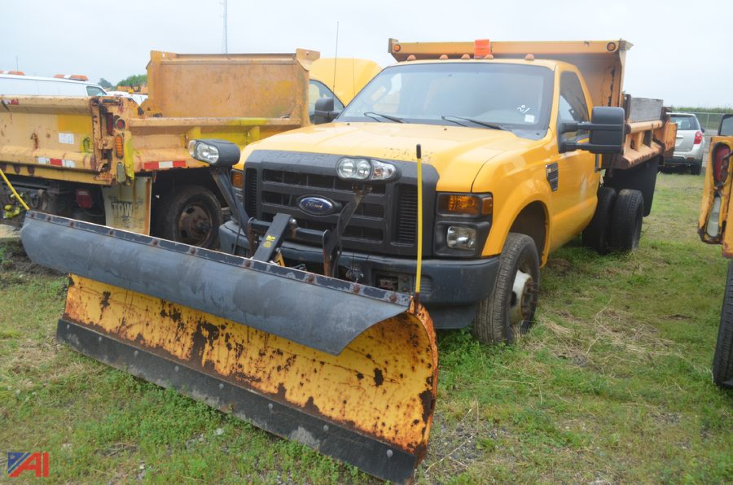Ford F-350 Super Duty dump truck with front snow plow attachment in foreground and yellow dump bed, daytime outdoor lot scene.