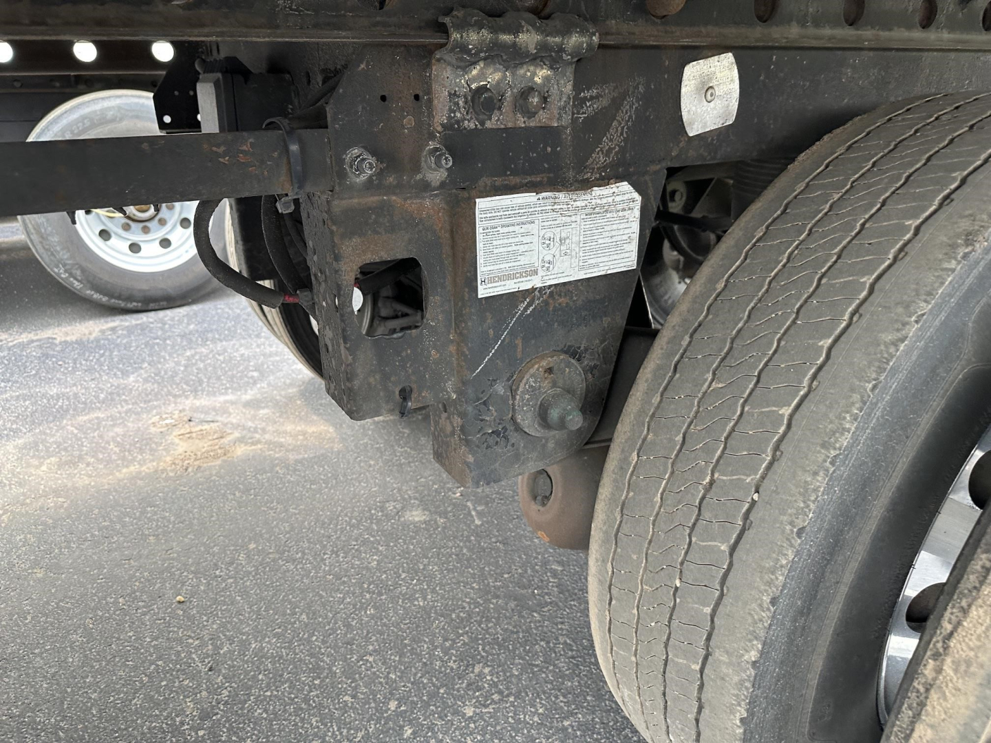 Trailer undercarriage showing frame rails, air brake line bracket, and a label on the seizing component near a single rear tire of a refrigerated trailer.