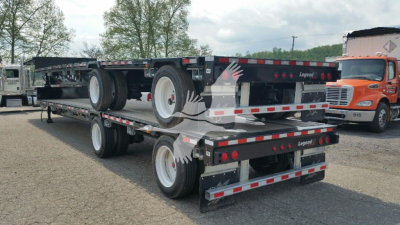 Manac drop deck trailer in a yard, dual axle rear view showing label and reflective strips, with another truck in background.