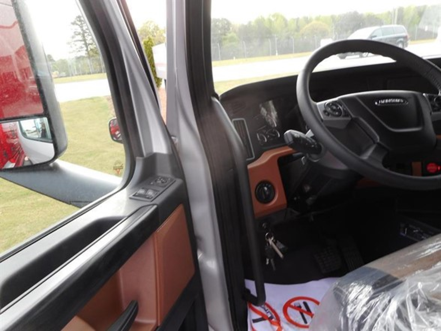 Truck cab interior view from driver's door showing steering wheel, dashboard, and orange door trim; plastic-wrapped seat and floor area visible.