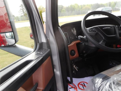 Truck cab interior view from driver's door showing steering wheel, dashboard, and orange door trim; plastic-wrapped seat and floor area visible.