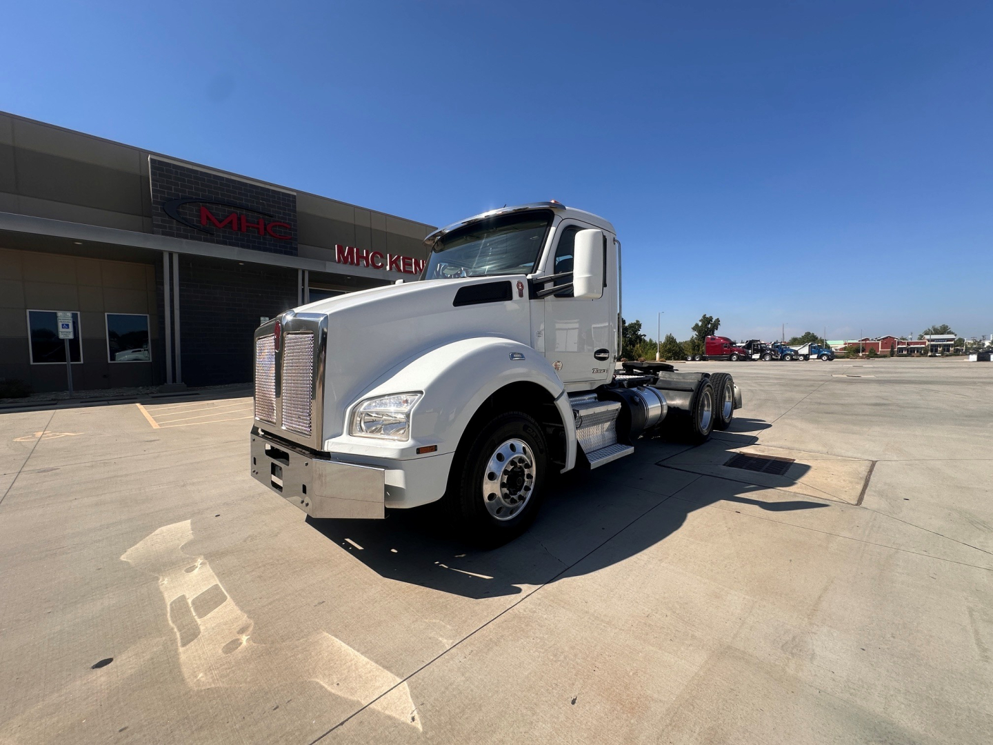 Kenworth T880 conventional day cab tractor in a dealership lot, side/front view, white cab with chrome grille, no trailer attached, clear sky.