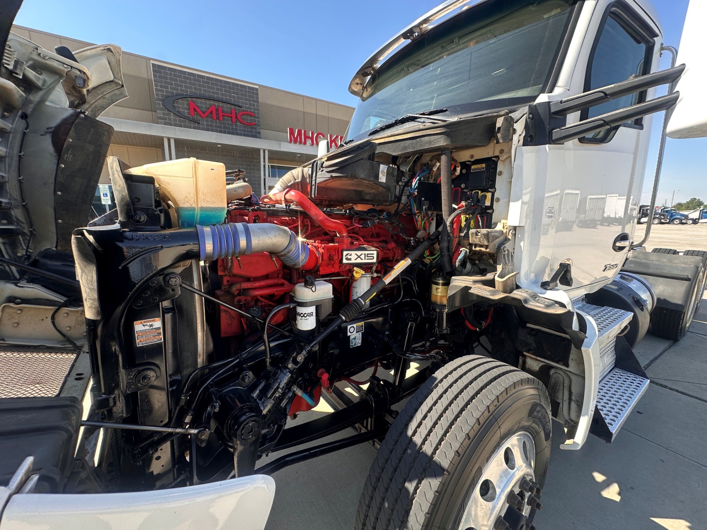 Kenworth T880 engine bay with open hood displaying red engine components, hoses, and wiring; side view shows front wheel and cab area.