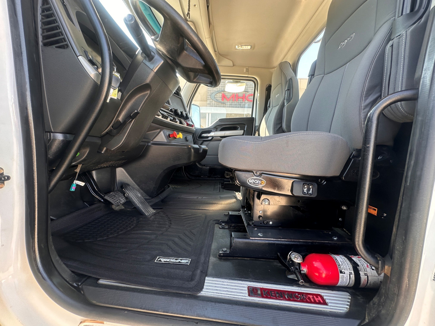 Kenworth T880 interior cab view showing driver seat and floor mat with Kenworth logo, steering column, and fire extinguisher mounted under seat.
