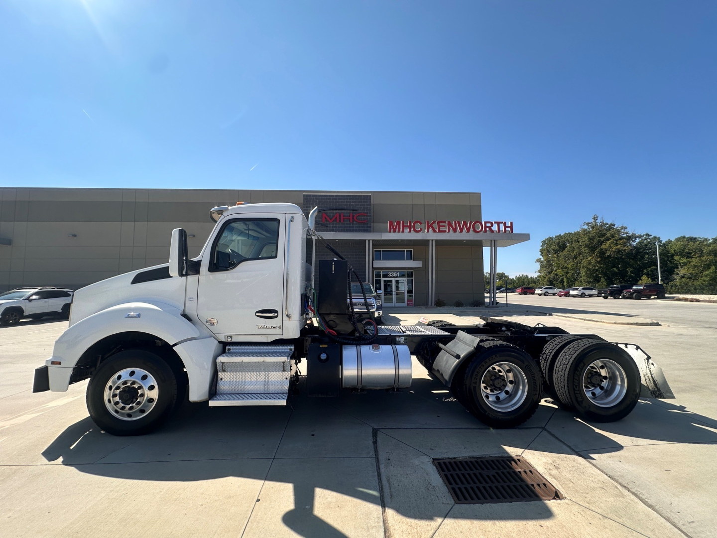 Kenworth T880 tractor in a dealership lot, side view showing chassis, fuel tanks, and three rear axles; no visible text on image aside from the dealership building in background.