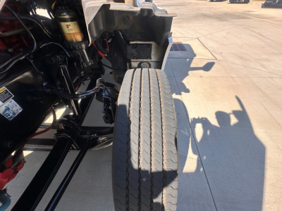 Kenworth T880 front wheel and chassis view showing dual rear-axle configuration, exposed suspension components, and part of the engine bay on a concrete surface.