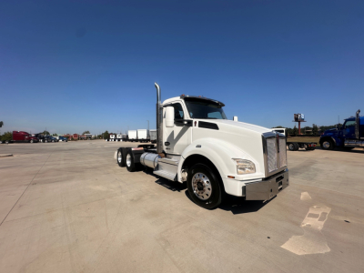 Kenworth T880 conventional daycab truck in white, front three-quarter view, showing V-shaped chrome grille, tall exhaust stacks, dual rear wheels, large fuel tanks.