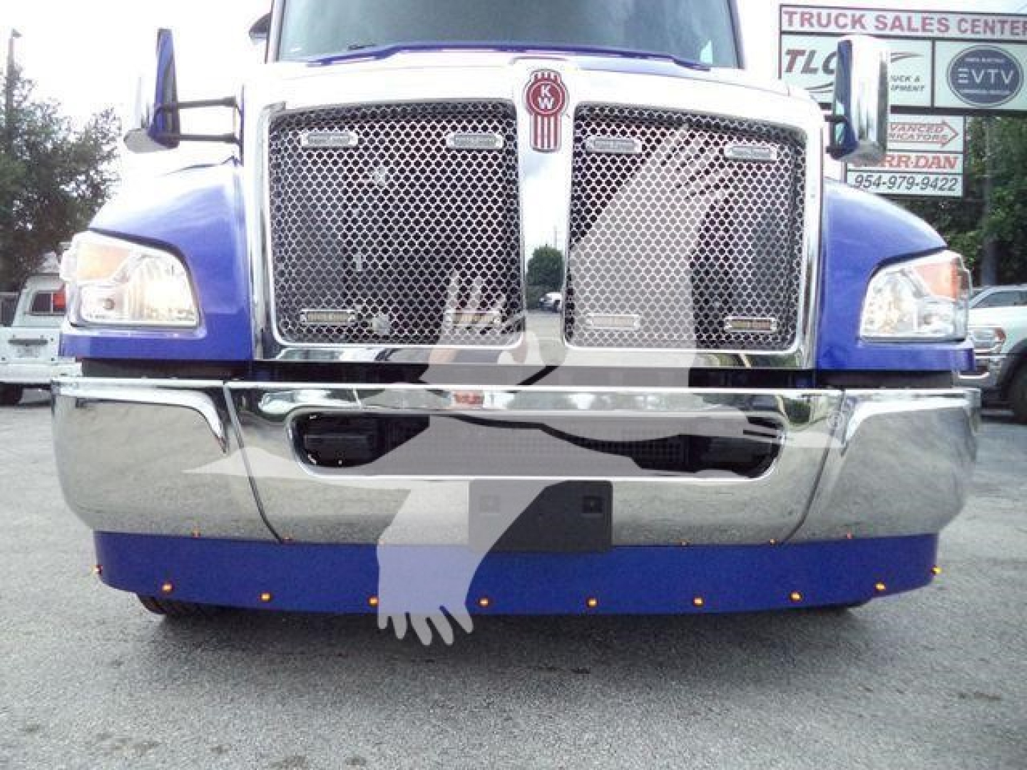 Kenworth T-head front view showing chrome grille, dual large bumper, and blue cab with emblem center; truck in a dealership lot.