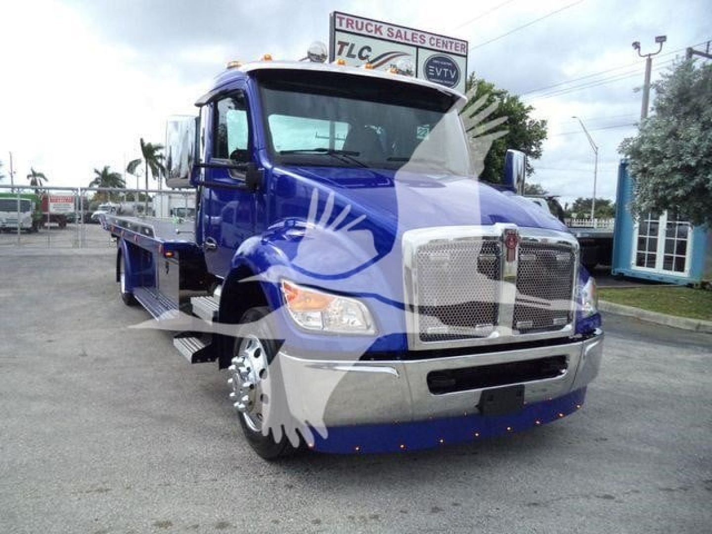 Blue Kenworth T280 rollback truck with chrome grille and side rails, showing front and left side; dealership signage in background.