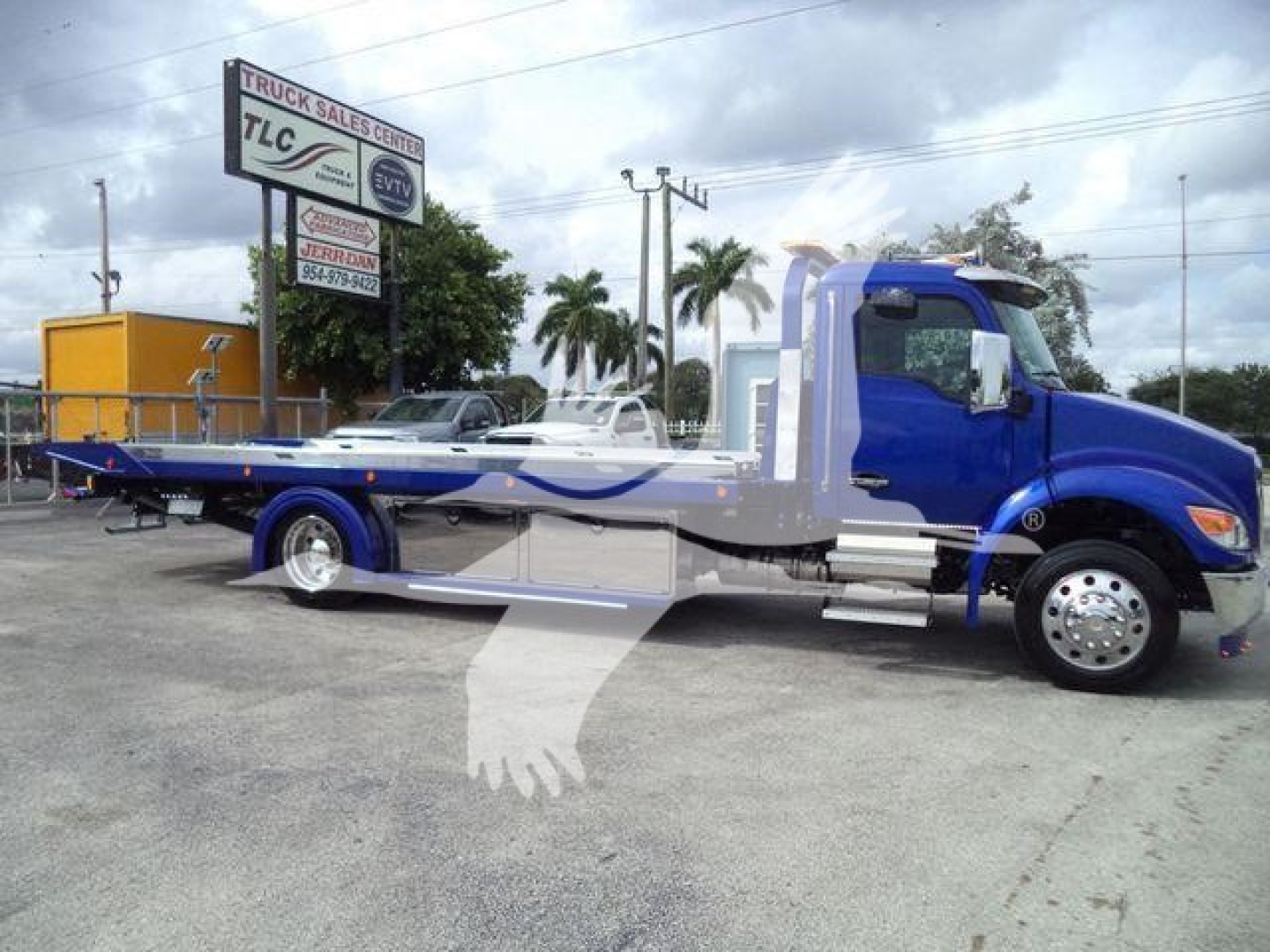 Blue Kenworth T280 rollback tow truck in a dealership lot with a long wheelbase, chrome exhaust, and tow bed visible.