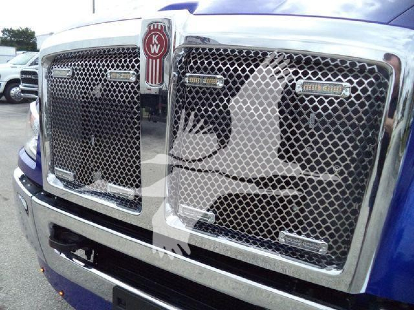 Front grille of a Kenworth truck showing chrome bumper and mesh grille with KW emblem and Peterbilt-like badges.