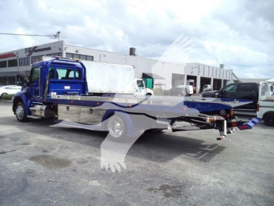 Kenworth tow truck/rollback chassis in blue with flatbed and winch, warehouse in background. Visible text: none legible.