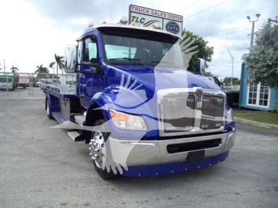 Blue Kenworth T280 rollback truck with chrome grille and side rails, showing front and left side; dealership signage in background.