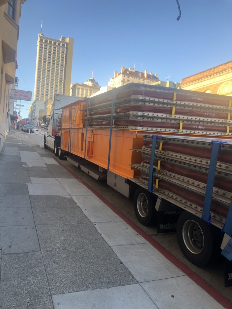 Trailer loaded with stacked metal decking and orange side panels, parked on city street, Big Bubba drop deck trailer by Reitnouer.