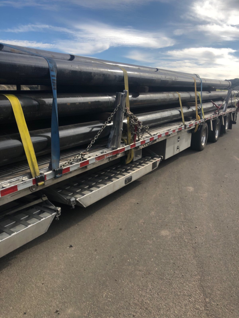 Trailer load of long steel pipes secured with blue and yellow straps on a drop deck trailer. Visible is multiple black pipes, metal side rails, and a walkway.