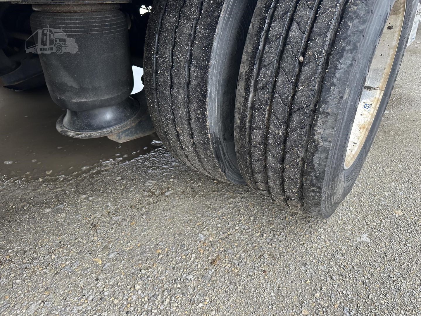 Trailer wheel and suspension area with dual tires; visible rubber tire treads and a black storage drum on the left; small white logo of a truck on the inner frame.