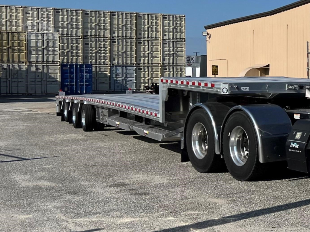 2019 Reitnouer Big Bubba drop deck trailer showing long flatbed with dual rear axles and stacked metal deck rails; background pallets and warehouse.