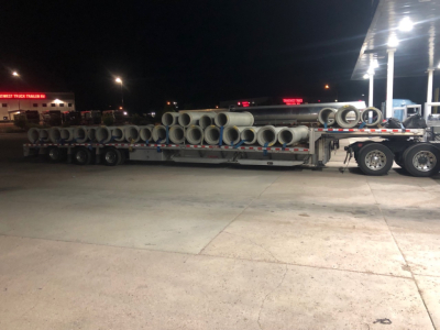 Flatbed trailer loaded with numerous metal pipes and cylinders, long trailer, nighttime scene at a truck stop.