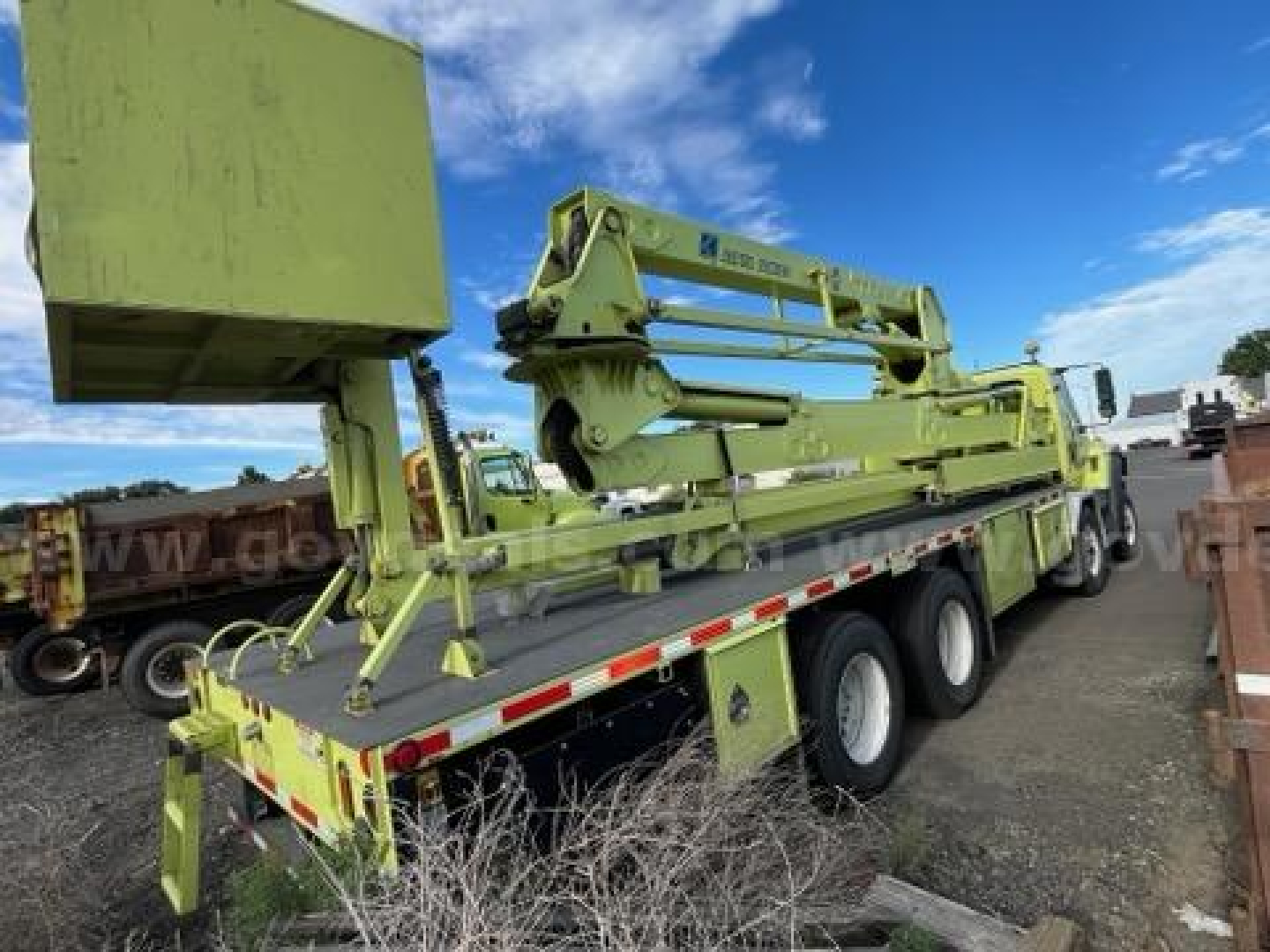 Green heavy equipment trailer with folded boom on a flatbed, showing hydraulic arms and stabilizers; some text on the boom reads 'RAYMOND' or similar. Wide trucks and blue sky in background.