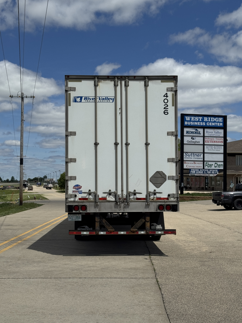 Rear view of a white Utility 4000D-X trailer with doors closed, showing Riverside Valley logo and rear bumper with taillights; road and building signage in background.