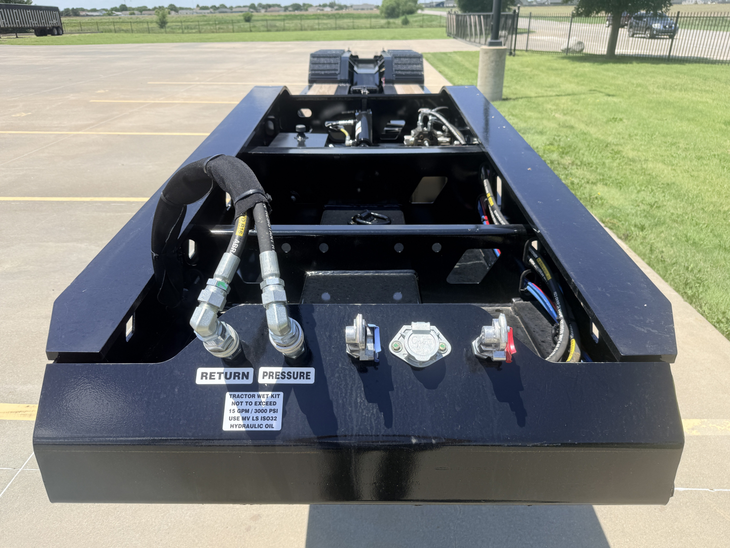 Trailer deck control area with hoses, hydraulic fittings, and pressure gauges; two metal caps and label reading RETURN PRESSURE visible in foreground.