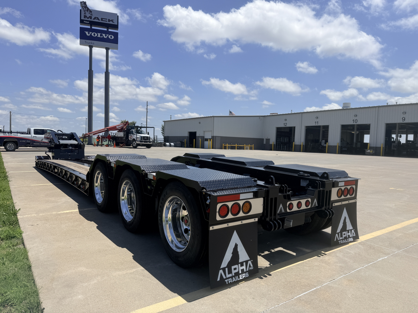 Alpha Trailers lowboy trailer with 18-inch LDH deck, dual rear axles, black frame, white Alpha mudflaps, shown from rear-left side in a dealership lot.