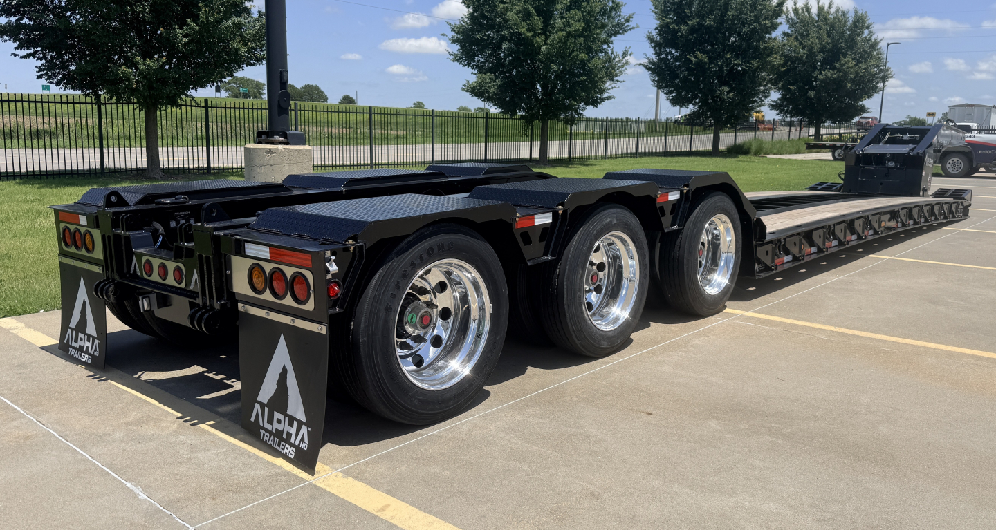 Lowboy trailer showing rear and side view with three axles and black deck. Alpha branding on mudflaps. 90-degree lights and chrome wheels visible.