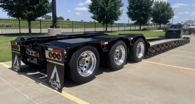 Lowboy trailer showing rear and side view with three axles and black deck. Alpha branding on mudflaps. 90-degree lights and chrome wheels visible.