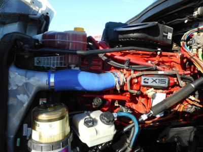 Freightliner Cascadia engine bay showing X15 engine with blue silicone hose, coolant reservoir, and various fluid reservoirs.