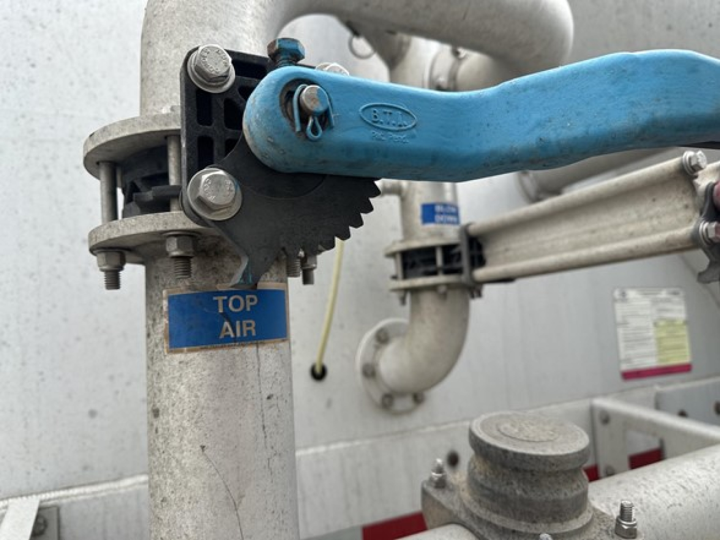 Pneumatic trailer top air clamp and blue lever on vertical silvery pipe; blue label shows TOP AIR with surrounding metal framework and piping.