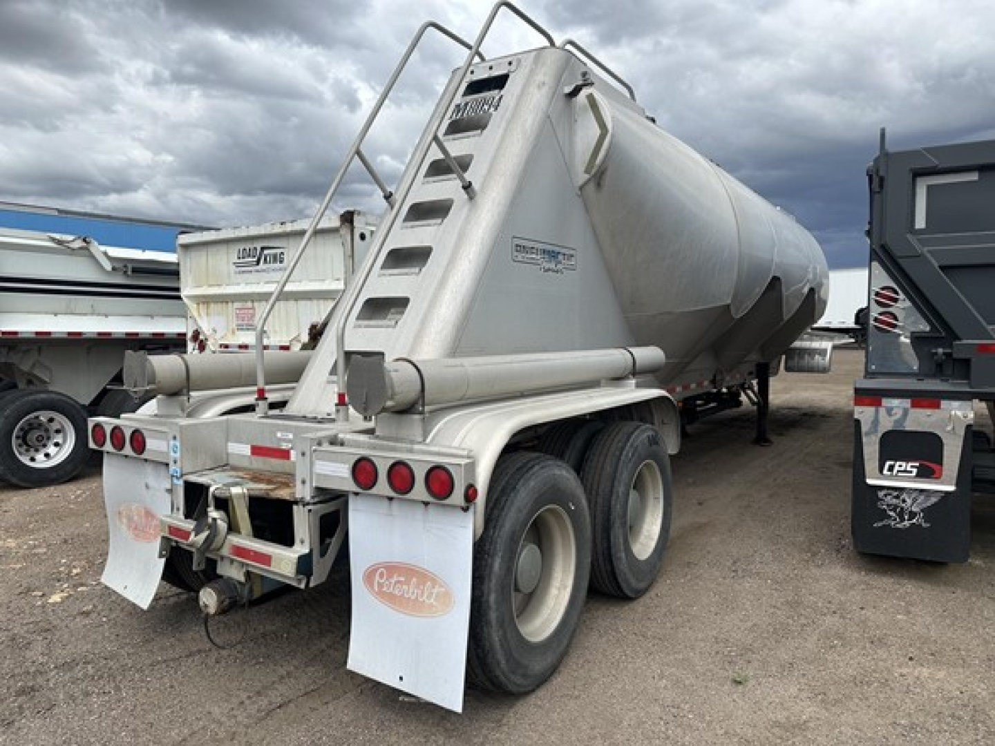 Trailer rear view of a Mac 1050 Cu Ft pneumatic dry bulk trailer showing rear lights, mud flaps, and ladder; tank with curved end and branding stickers visible.