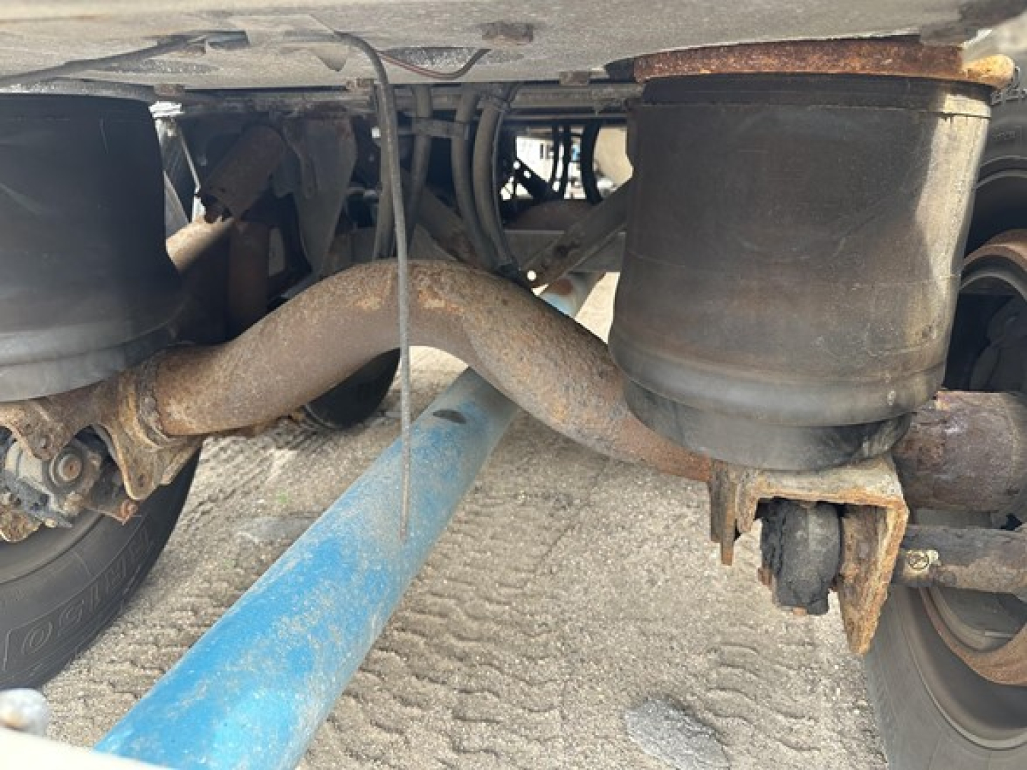 Trailer underside showing air suspension bags and exhaust-like pipe with rust; wheel hubs and leaf spring visible.