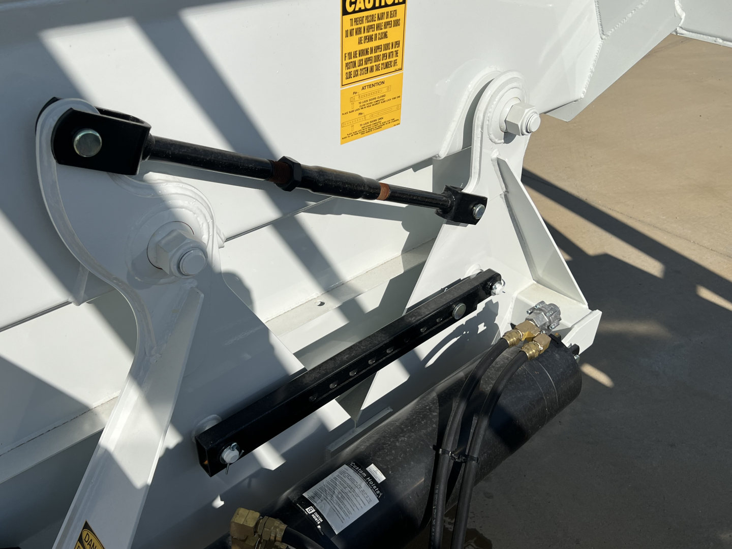 Dump trailer rear frame with hydraulic air ride linkage and warning label, white paint, black support arms and hoses visible.